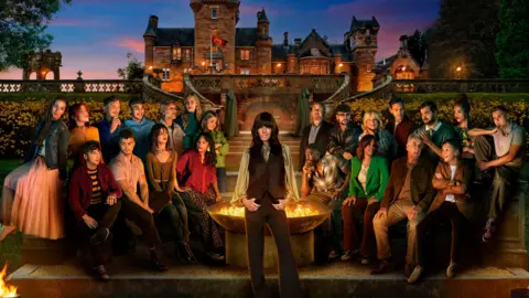 BBC/PA The contestants taking part in this year's The Traitors sit outside the Castle at night time near a large fire pit. Claudia Winkleman is standing in front of the fire place looking directly at the camera. In the background is a large castle against the setting sun.