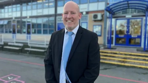 Headteacher Jon Kirby smiling for a picture in front of the school. He is wearing a dark suit and a blue tie.