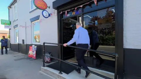 A man wearing a blue shirt and dark trousers walks up a concrete wheelchair-accessible ramp to the large black-framed glass doorway of a village shop. The brick building is painted light-grey and black. Signs advertise a Post Office, the National Lottery and ice creams. A man stands to the left wearing a blue jacket.
