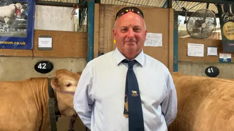 A man wearing a white shirt and a navy blue tie stands in front of several blonde cows.
