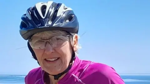 Pat Marshall, who is standing at the coast with her bicycle, is wearing a blue helmet, glasses and a pink top. She is looking at the camera and smiling slightly.