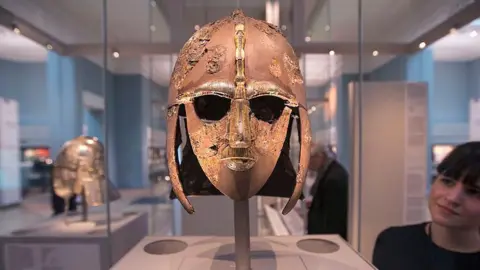 Oli Scarff/Getty Images The Sutton Hoo helmet in a glass case at the British Museum with a lady looking at the item from behind. The helmet is made from iron and copper and covers the entire face except the eyes. 