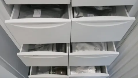 The National Botanical Garden of Wales A set of white laboratory drawers pulled open to reveal neatly stacked silver foil packets. Each packet contains stored seeds, labelled and organised for preservation.