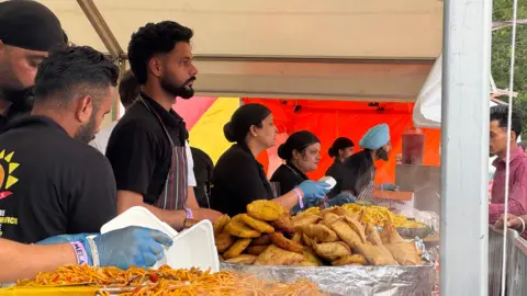 Staff at a food stall serving up a variety of foods from large silver dishes, including samosas.