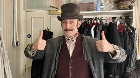 Jamie Tyler Jamie Tyler wearing a burgundy waistcoat, a tattered grey jacket, a striped shirt, a neck scarf and a top hat, and a moustache. He is giving the thumbs up. He is in a dressing room, and there are costumes on racks behind him.