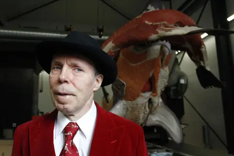 Getty Images Gunther von Hagens, wearing a red jacket and tie with white shirt, and a black hat, stands in front of an exhibit in Guben, Germany in 2016.