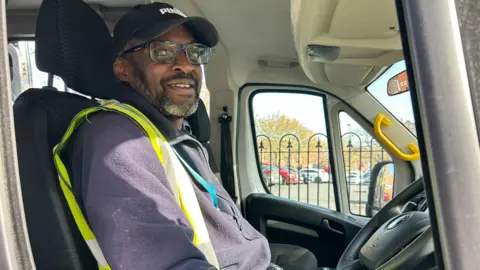 Gavin Kermack / BBC A man with a short salt-and-pepper beard and wearing glasses, a black Puma baseball cap and a purple fleece under a hi-vis waistcoat sits in the driving seat of a minibus.