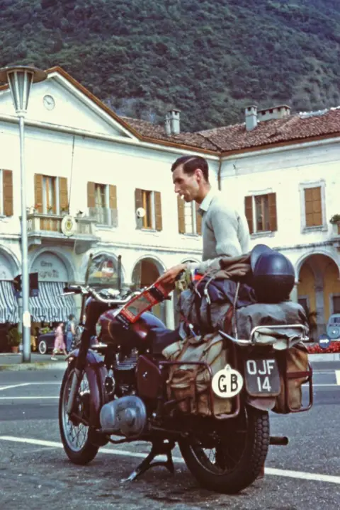 Ray Stokes Ray Stokes with his old motorbike with a GB sign on the back, and buildings and a hillside in the background