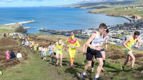 DAVE KNEEN A trail of runners running up Peel Hill, you can see the bay in the background, One person has a yellow chicken costume on.