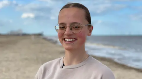 Mary Catchpole, smiling with her hair tied back and wearing glasses. She is standing on a beach near her home in Norfolk