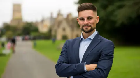 Joe Harris wearing a light blue collared shirt underneath a navy blue blazer. He is standing with his arms folded in front of a large green park area with historical brick buildings in the background. He is smiling at the camera, and has dark hair which is shaven short on the sides and a goatee style beard.
