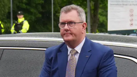 Donaldson has mostly grey hair and has exited a car. He is wearing a blue suit jacket, white shirt and speckled tie. He is also wearing glasses. A car is in the background as is a police officer.