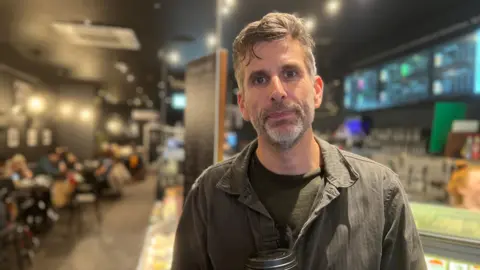 BBC A man with a trimmed slightly grey beard looking at the camera . He is wearing a black T-shirt with a dark green shirt on top and holding a coffee cup. He is standing in a coffee shop which is out of focus behind him.