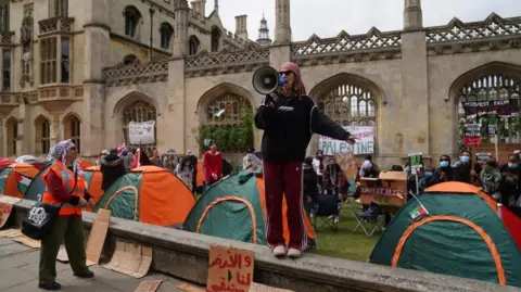 PA Media Protest in Cambridge: a woman wearing black top and maroon tracksuit bottoms stands on a wall holding a megaphone. A number of people are standing behind her on a grass area on which green and orange tents are pitched.