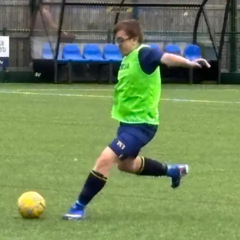 Supplied Taylor wearing a bright green training bib strikes a yellow ball on an artificial turf pitch. Team benches and perimeter fencing are visible behind him.