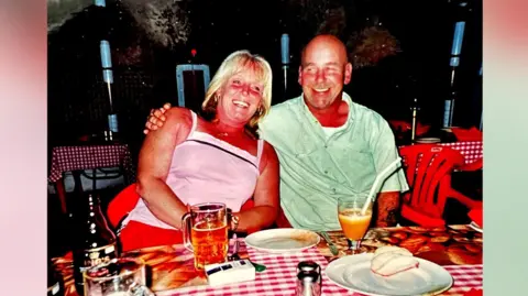 Diane Adams A man and a woman are sat in a restaurant while on holiday. They are both smiling and the man has his arm around the woman. They appear to be in their mid-50s. The table in front of them has several drinks on it and a plate of food.