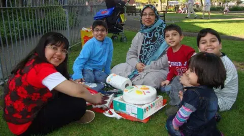 Dr Sabah Usmani, who has glasses and a headscarf, sits on the grass in a park, surrounded by five children, who are all smiling. In front of them is an uncut cake. A pushchair can be seen behind them.