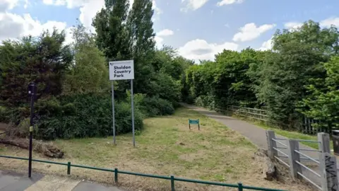 An area of green grass behind a small green metal fence. There are large green trees and bushes with a concrete path. On the grass there is a tall sign that reads "Sheldon Country Park"