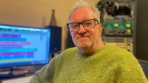 Simon Thake A man with white hair, black rimmed glasses and a fluffy green jumper sits in front of a computer screen with audio files