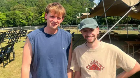 Jack on the left is wearing a short-sleeved dark blue T-shirt and has ginger hair. Luke is wearing a faded green cap with a beige T-shirt. They are stood on grass with chairs in rows behind them.