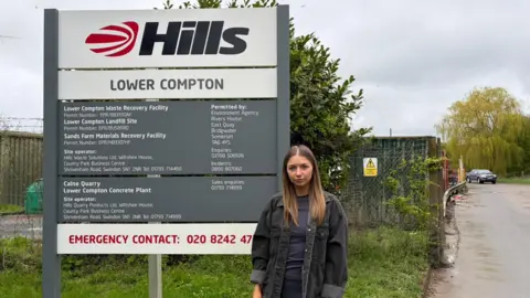 Resident Simone Partner is wearing a charcoal-grey shacket, standing in front of the Lower Compton landfill entrance gateway, with a large grey and white sign behind her with the Hills logo on it.
