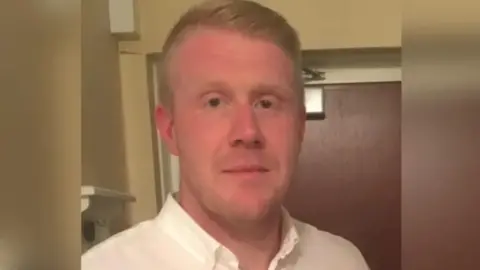 A clean-shaved man in a light-coloured shirt, with short, blond hair. A brown door is behind him. 