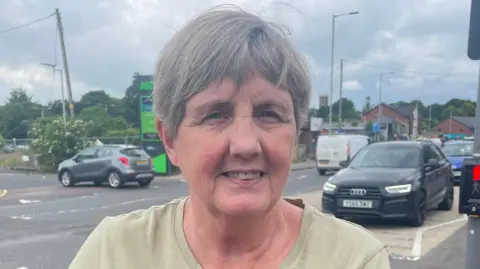 A woman, with short grey hair, wearing a light green t-shirt. She is standing on the side of the road.