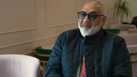An image of councillor Sidat, he is sitting in a cafe, with a white panelled wall behind him, next to him is an empty white chair. He is a bald man with a grey beard and is wearing dark glasses. He has on a padded blue jacket and a red tie.