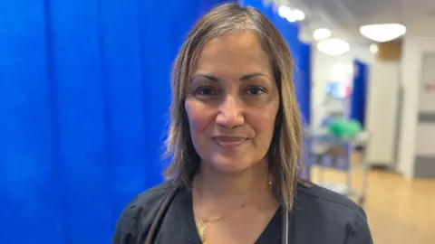 Dr Shewli Rahman stands in the resuscitation area, where the sickest patients are treated. She has near shoulder-length hair and has dark blue scrubs on with a stethoscope round her neck.