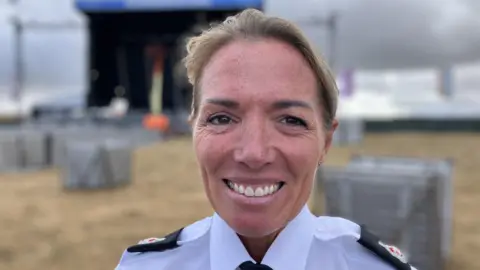 Vicky Howell, a woman with short pulled back hair, is smiling at the camera. She has police superintendent epaulettes on her shoulders and is wearing a white shirt with black tie