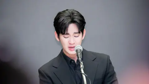 A tearful Kim Soo-hyun speaking into a microphone. He is wearing a fully black outfit.