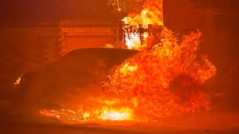 Facebook A car engulfed in bright orange flames. It is parked outside a property with a brick wall. The flames have spread from the car to a fence.