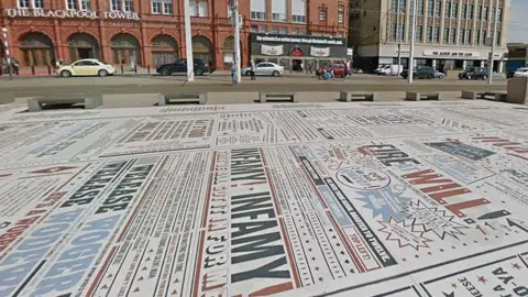 Street view image of the Comedy Carpet. A huge flat installation on the promenade full of text written in different fonts and sizes in red black and blue on a white background