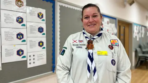 Evelyn smiles at the camera in her Scouts uniform. She has several badges on her cream coloured shirt with a blue and white neckerchief. Evelyn has dark hair which is tied back into a ponytail.