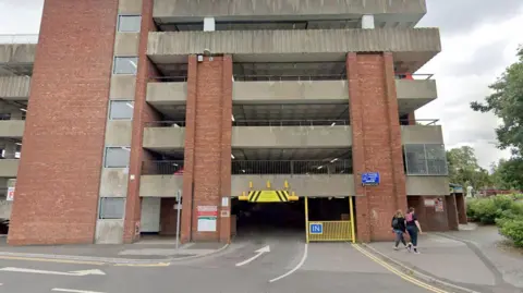 Google Maps The front of a multi-storey car park, made up of red brick and grey concrete. It has five upper levels and the entrance has a blue sign saying 'IN'.