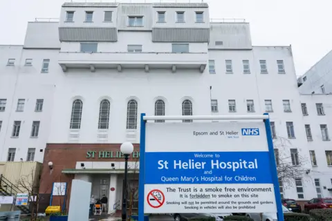 Exterior of St Helier Hospital shows white building entrance with blue and white Epsom and St Helier NHS trust sign welcoming patients and stating it is smoke free