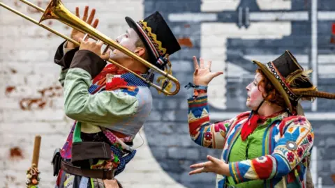 SO Festival A man and a woman wearing colourful clothes, hats and face paint perform in a street in front of a brick wall painted white and blue. The man is playing a trombone while the woman is dancing.