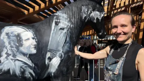 Andrew Turner/BBC Lois Cordelia smiling, next to her artwork; a black horse painted with images of Black Beauty and author Anna Sewell.