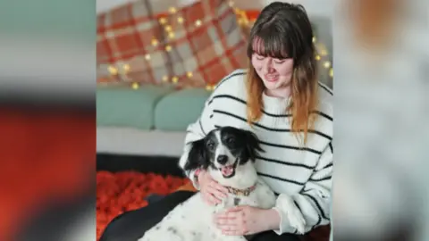RSPCA Floss, a black and white springer spaniel, is hugged by her owner Shannon Hamilton in a living room. Shannon wears a black and white striped jumper, and has long brown hair with lighter ends.