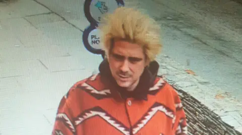 Alexander Key is walking in a street. He has peroxide blonde hair and a mustache. He is wearing a bright red coat which has a bold zigzag print in white and black.
