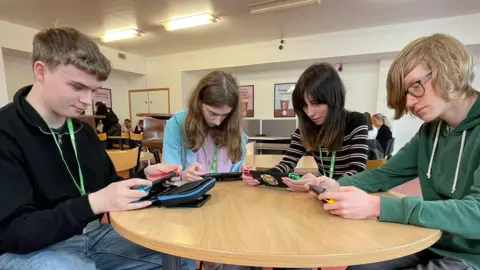 Laura Foster/BBC News Four students are sitting at a wooden round table at the college coffee shop, each looking down playing a different game on their Switch consoles.