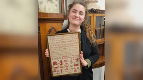 Auctioneer holding the sampler. It sits in a brown portrait frame, and is around 2 feet long and a foot wide. The stitching is red and shows various fonts of stitched letters from A to Z as well as flower pattern motifs and images, including a house and a steam engine.
