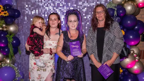 A lady with purple braided long hair wearing a black and purple dress poses smiling alongside two other women, one holding a young girl in a red dress