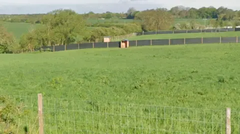 LDRS A field in Wragby, which has fences around and a small shed on one side. There are hills and trees in the background. 