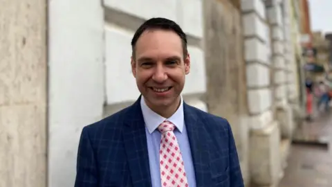 Laura Coffey/BBC Dan Lister with short dark hair wearing a blue jacket over a blue shirt, and a red and white tie. He is standing in front of a stone wall.