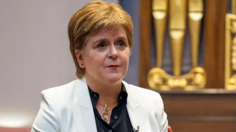 Nicola Sturgeon wearing a white jacket and black shirt holding her hands out in front of her as she talks 