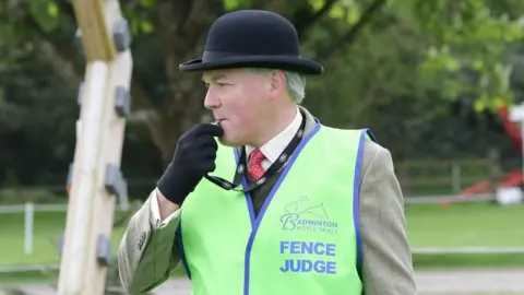 Martin wears a high-vis fence judge jacket and a bowler hat. He is blowing a whistle.
