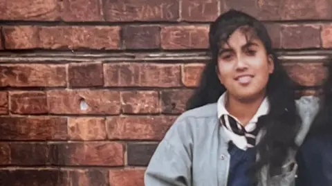 Rahena Begum Teenage girl in school uniform smiles at the camera, leaning against a brick wall.