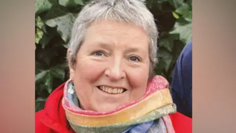 Westmorland and Furness Council Councillor Judith Derbyshire. She has short grey hair and is wearing a red coat and colourful striped scarf. She is outdoors, stood in front of a leafy bush.