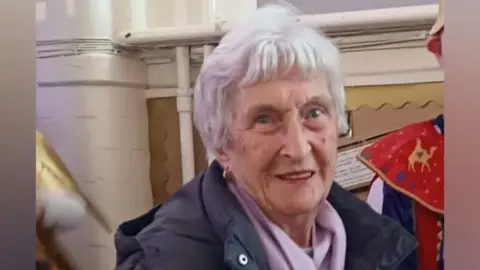 Rose Allan is looking just past the camera and smiling slightly. She has short white hair and is wearing a black coat and a lilac scarf.